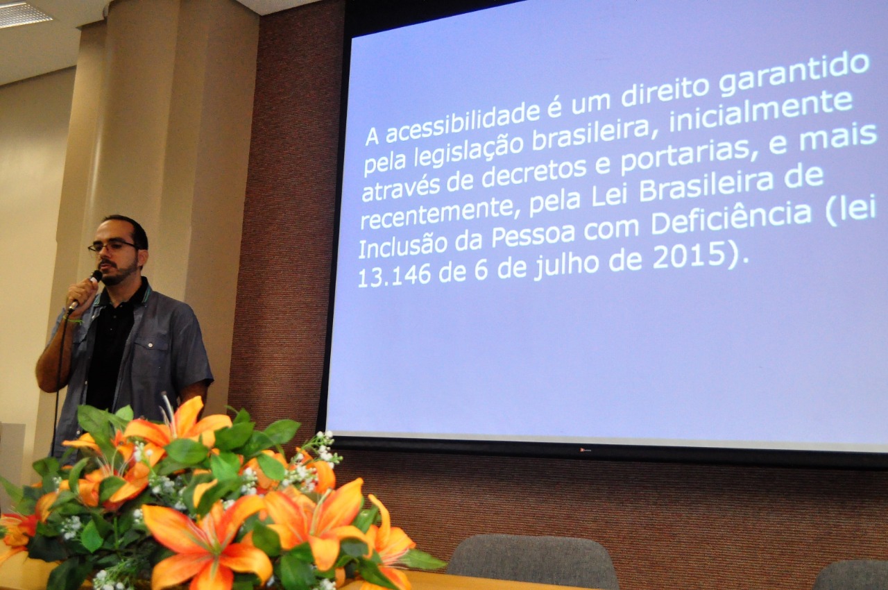 Fotografia de Jorge ao microfone. Ao fundo, uma projeção com letras brancas e fundo azul, com texto sobre a legislação de acessibilidade no brasil