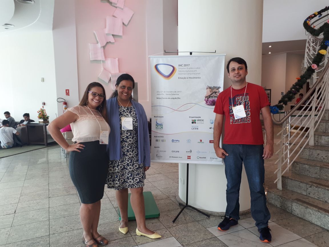 Priscyla, Carolina e Bruno em frente ao banner do evento, no hall de entrada